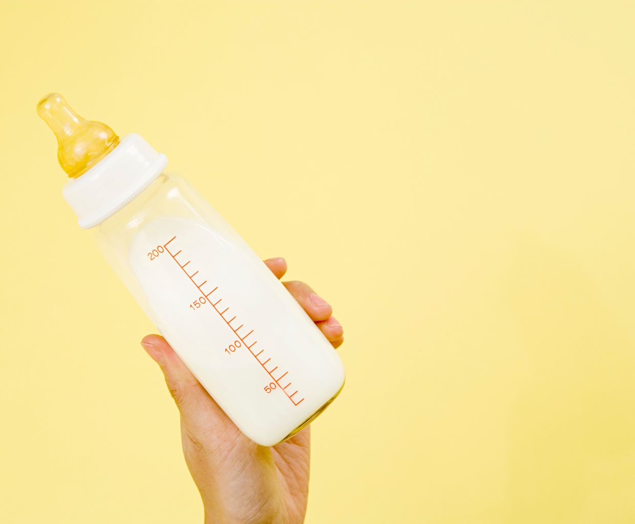 Hand holding a baby bottle filled with milk against a pale yellow background