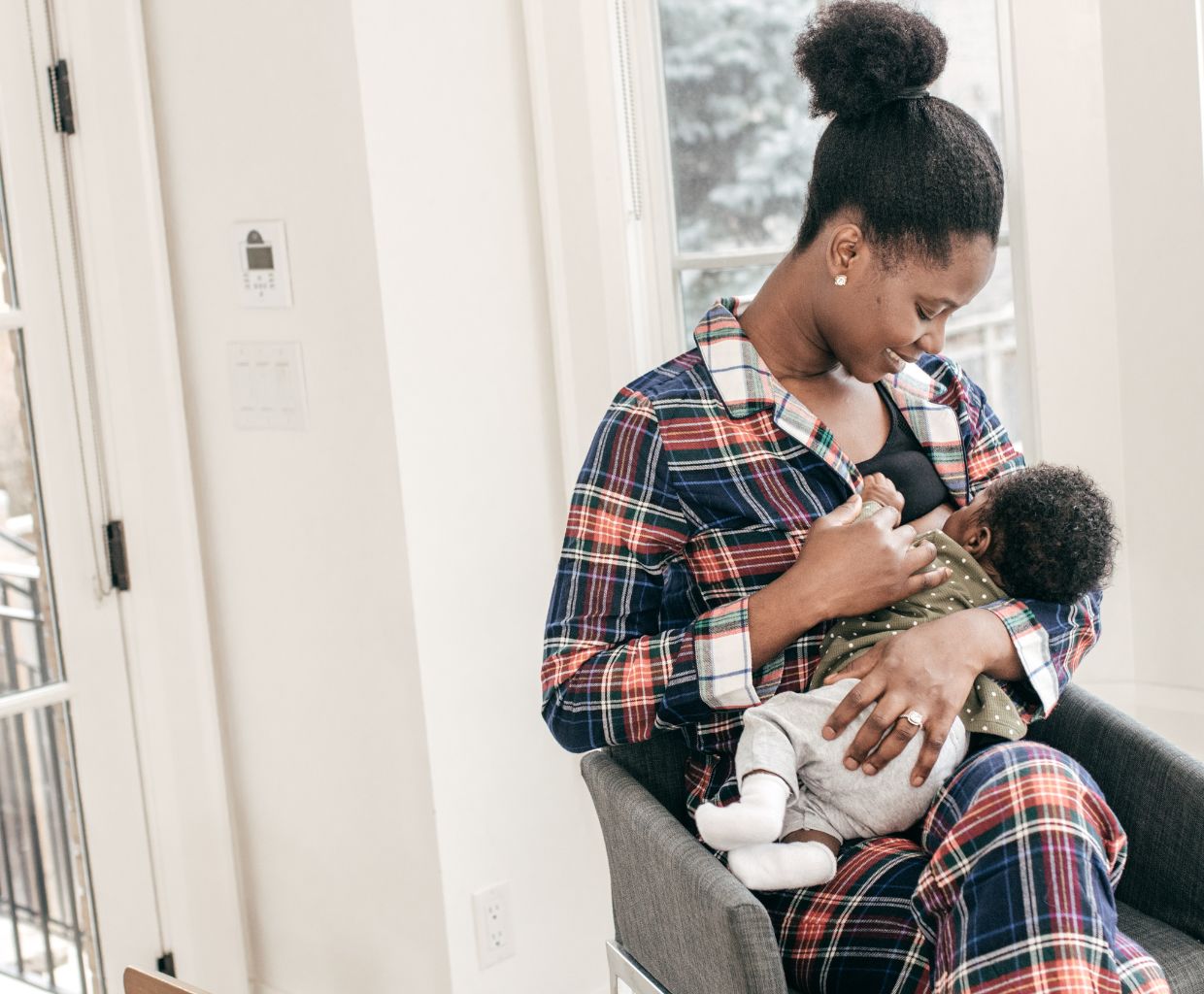 Smiling mother in plaid pajamas breastfeeding her baby in an armchair by a window