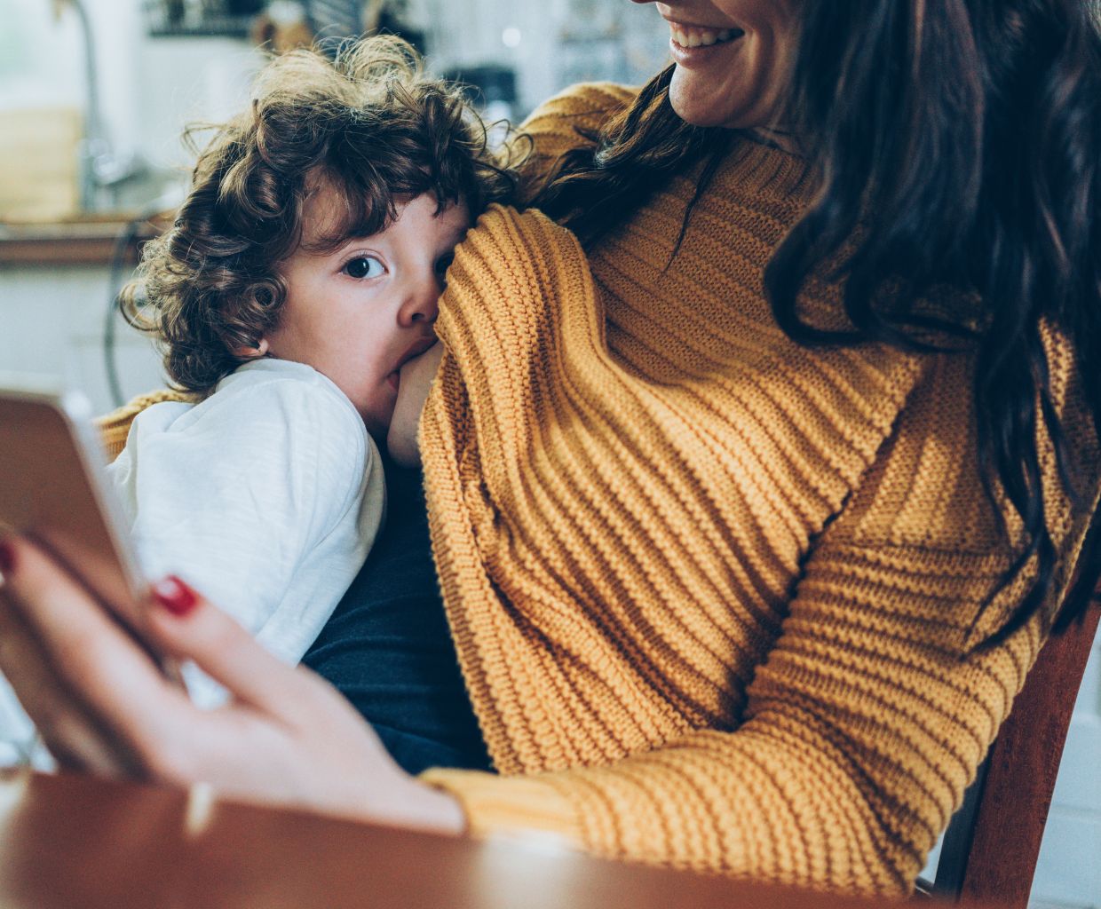 Curly-haired toddler breastfeeding on a smiling mother's lap at a kitchen table