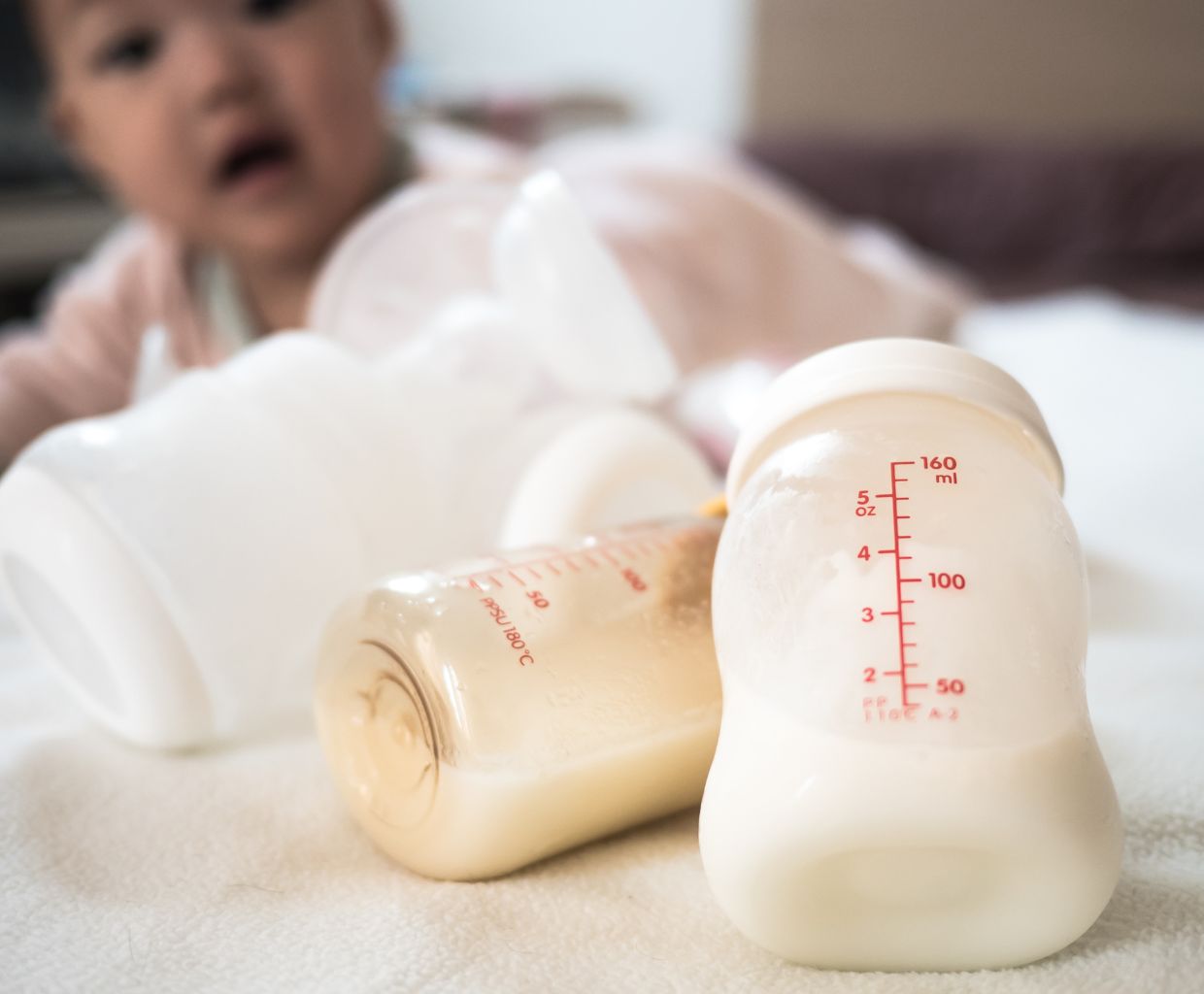 Two bottles of expressed breast milk standing on a soft blanket with a baby blurred in the background