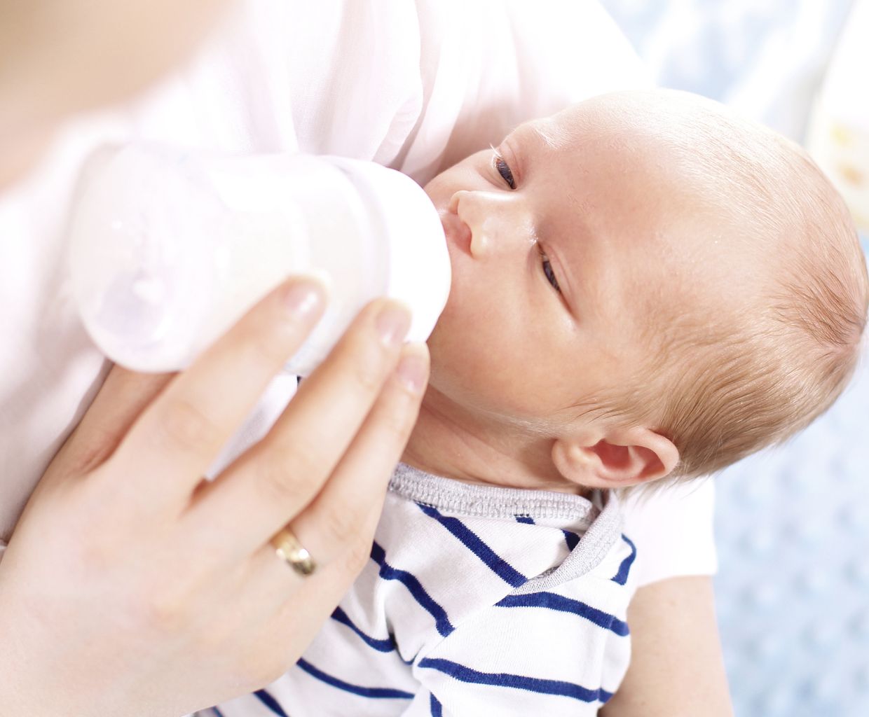 Newborn baby in a striped onesie drinking warm breast milk from a bottle held by a parent
