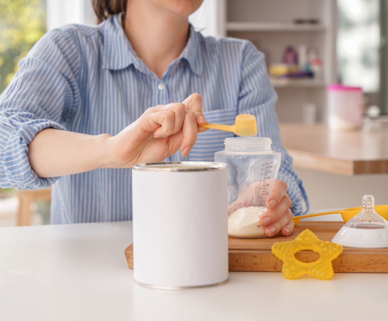 Parent scooping powdered infant formula into a baby bottle on a kitchen table