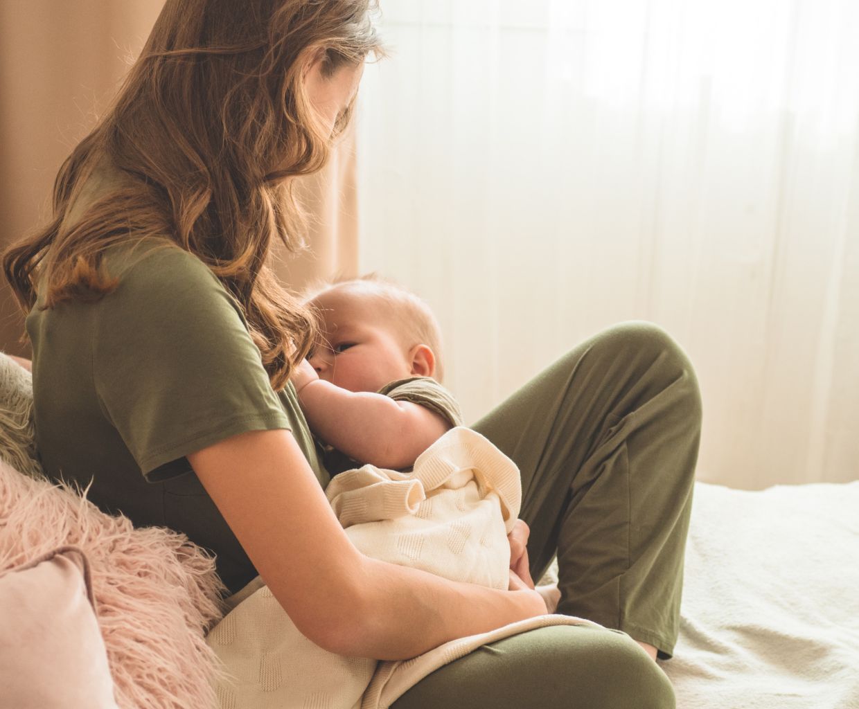 Woman with long hair sitting on a bed breastfeeding a baby wrapped in a cream blanket